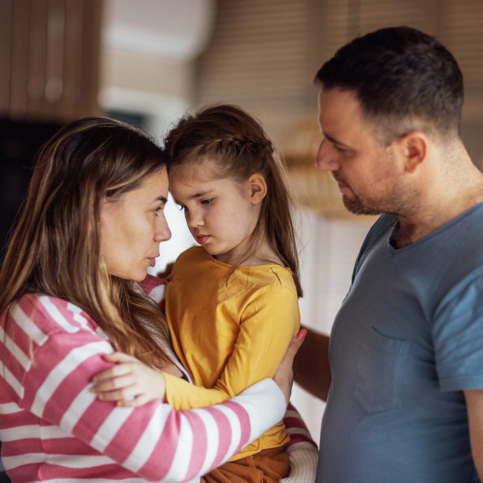 Young parents comfort their cute daughter. Beautiful woman hugs and talks to little girl in living room at home. Dad and mom take care of their child. Tender moment family support and understanding.